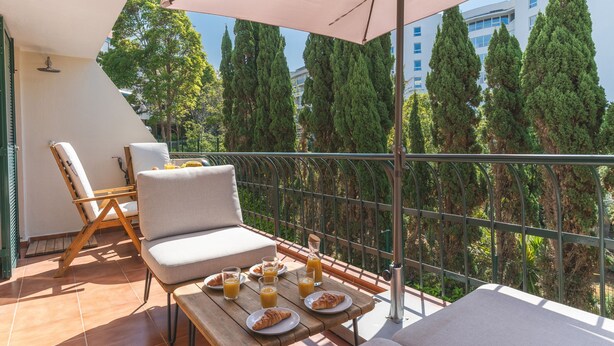 A balcony with a table set for breakfast, a sofa, and a chair, overlooking a row of tall trees and a building. - Costa Azul by Atlantic Holiday (Funchal)