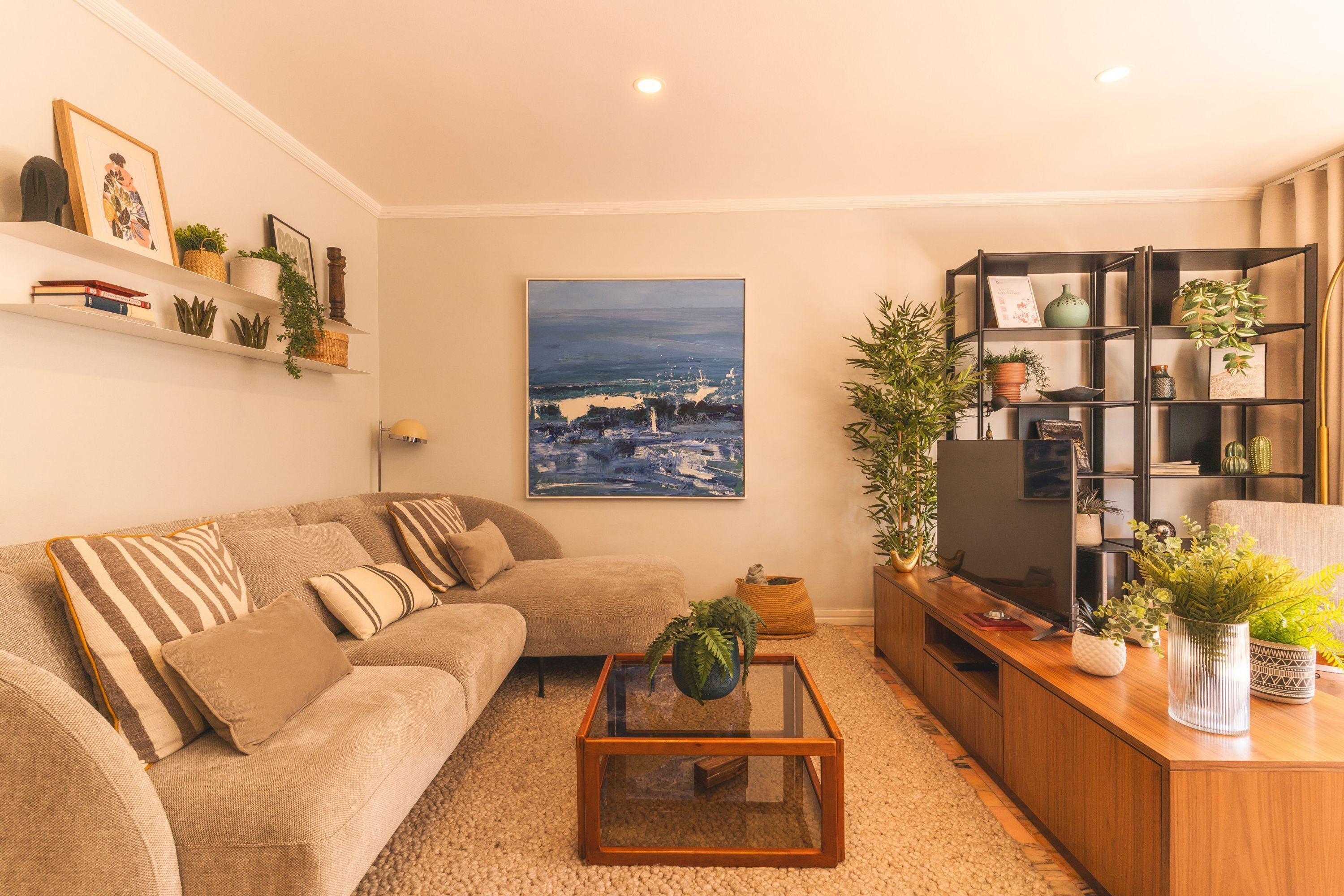 A living room with a sofa, coffee table, TV stand, and shelves with plants and books.