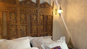 Un lit avec du linge de lit blanc et un seul coussin décoratif, une tête de lit en bois ornée de motifs complexes et un luminaire fixé au mur.