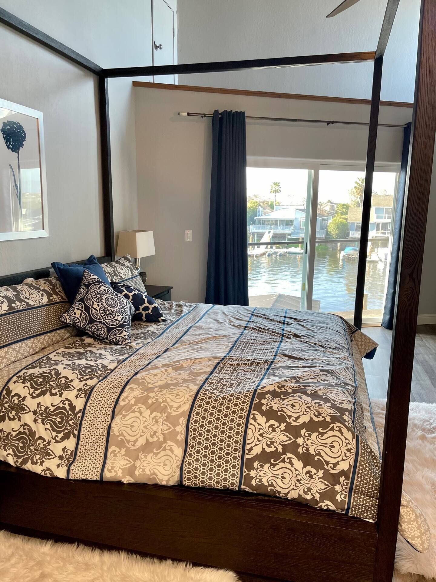 A four-poster bed with patterned bedding, a nightstand with a lamp, and a view of a body of water through large windows.