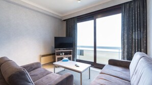 A modern hotel room with two sofas, a TV on a stand, a small table, and a large window with curtains.