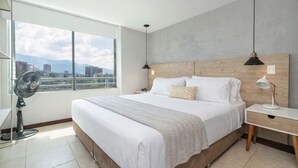 A modern hotel room with a large bed, a nightstand, a desk, a fan, and a view of the cityscape through large windows. - Penthouse Apt Poblado, City View, Rooftop Jacuzzis (Medellín)