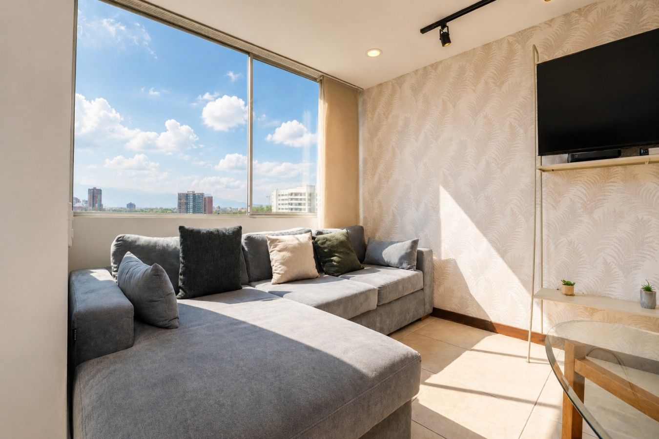 Ein modernes Wohnzimmer mit einem grauen Ecksofa, einem flachbildfernseher an der Wand und einem großen Fenster mit Blick auf die Stadt.