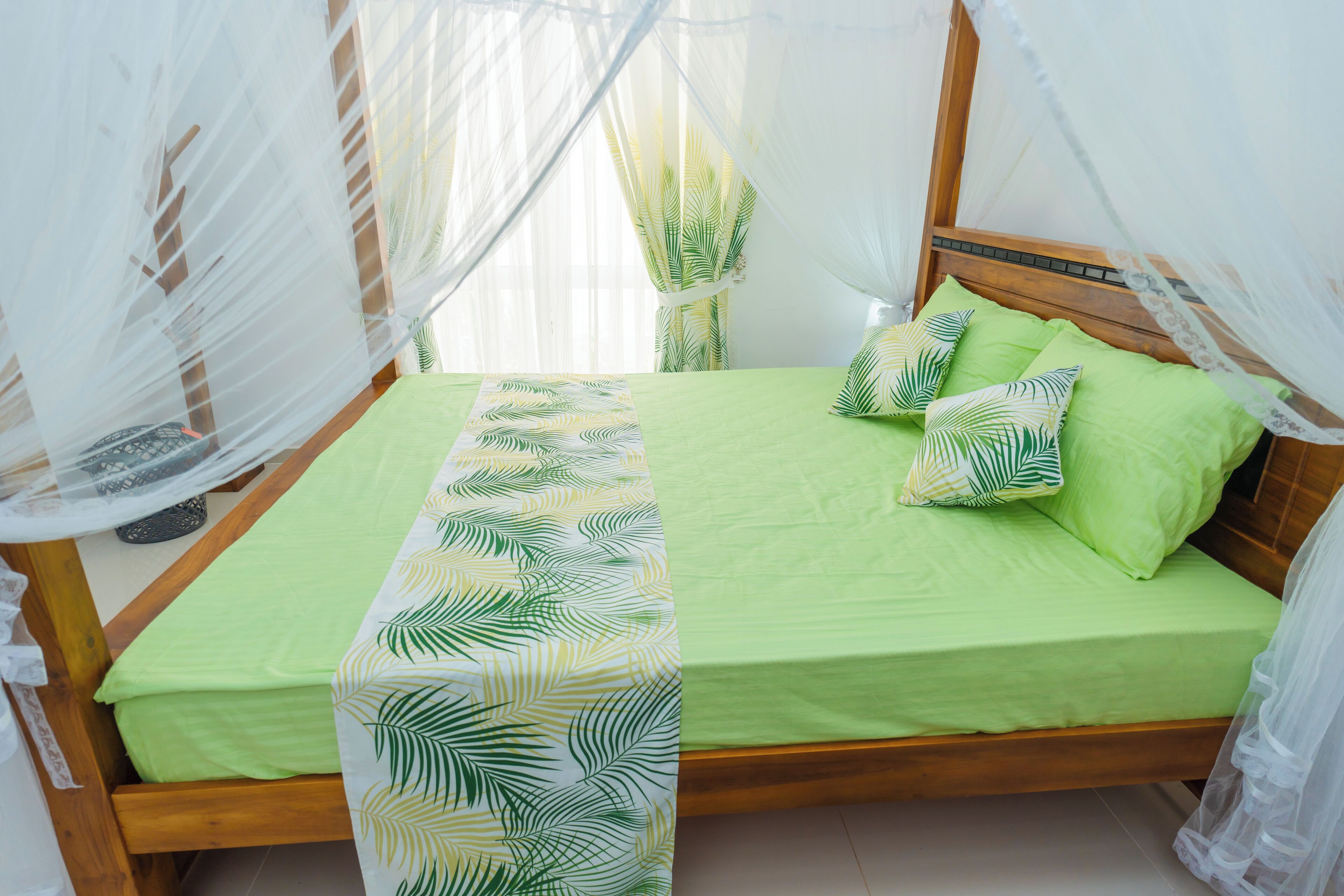 Un lit avec une couvre-lit verte et un chemin de lit orné de motifs de feuilles, une tête de lit en bois et un baldaquin en tissu blanc transparent.