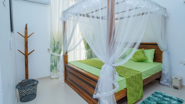 Une chambre avec un lit en bois, une literie verte, un baldaquin blanc, un porte-manteau en bois, une plante verte et une petite poubelle.