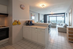 A modern kitchen with white cabinets, a stove, and a coffee maker. There is a dining area with a white table and chairs, and a view of the ocean through large windows. - Apartment in Middelkerke Near the Beach (Middelkerke)