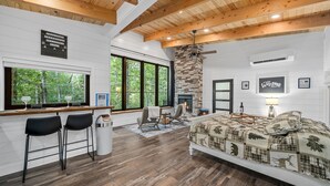 Une chambre moderne avec une cheminée en pierre, un coin bar avec des tabourets et un grand lit agrémenté d’une couvre-lit sur le thème de la forêt.