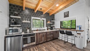 A modern kitchen with a stone backsplash, wooden ceiling beams, stainless steel appliances, and a bar area with stools. - Treehouse - Twilight (Jonesburg)