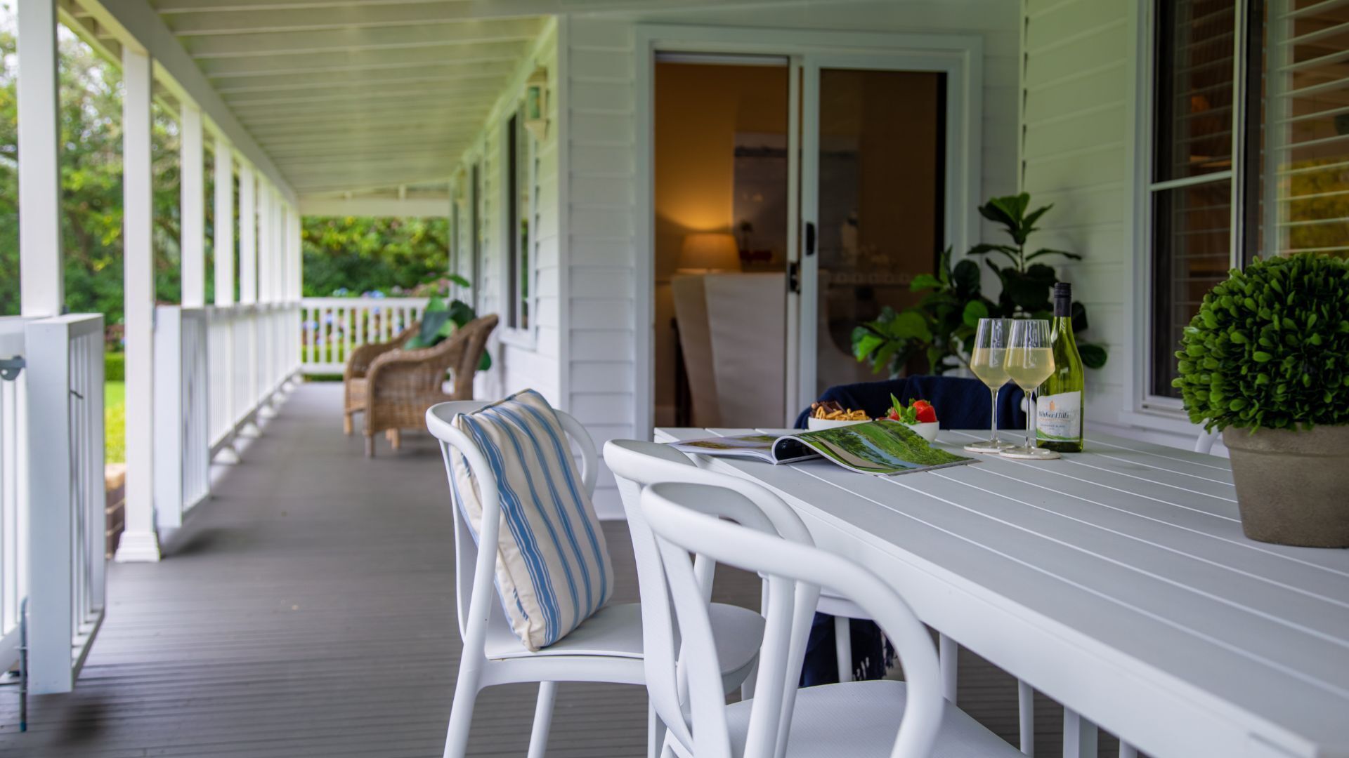A covered porch with white furniture, a table set with a menu, wine glasses, and a bottle.