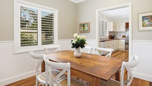 Een eetgedeelte met een houten tafel en witte stoelen, een bloevas en uitzicht op de keuken en groen buiten door de horizontale zonwering.