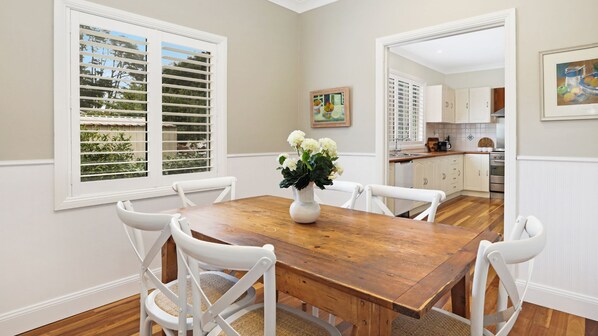 Un coin repas avec une table en bois et des chaises blanches, un vase de fleurs, et une vue sur la cuisine et la verdure extérieure, visible à travers les stores de la fenêtre.