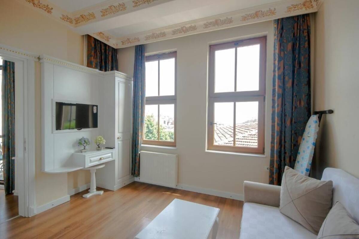 A room with a white sofa, a small white table, a TV mounted on the wall, and two windows with curtains.