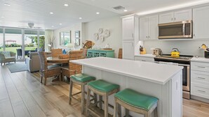 Una cocina moderna con una isla central, armarios blancos y un comedor con mobiliario de mimbre.