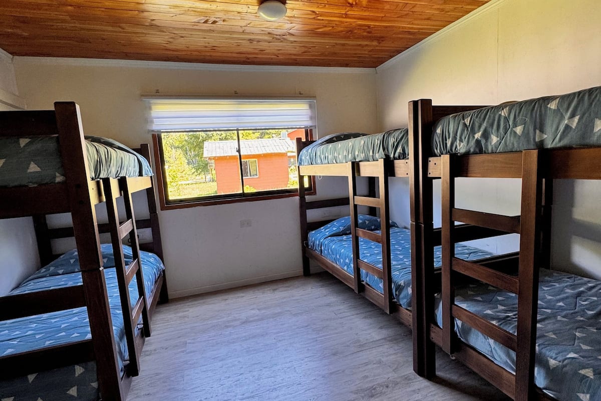 A room with four bunk beds arranged in a corner, wooden ceiling, and a window with a view of a building outside.