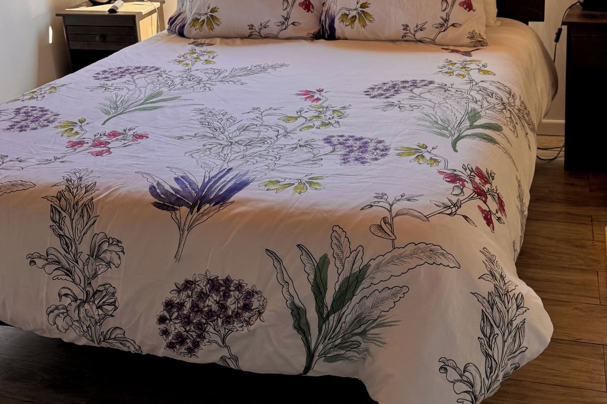 A bedroom with a large bed, floral bedding, a nightstand, a lamp, and a window with a view of trees.
