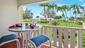 A balcony with a table set for two, overlooking a well-manicured lawn and palm trees, with a view of the ocean and a building in the distance. - Harbour Heights #36 Seven Mile Beach, 2 Bedroom, 2.5 Bathroom Second Floor Townhouse (Seven Mile Beach)