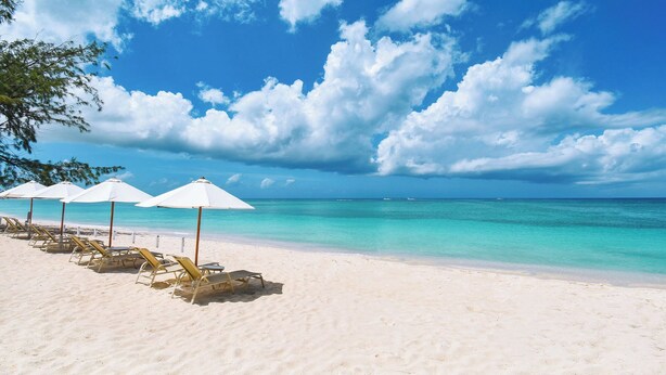 Een strand met wit zand, helder turquoise water en een rij witte strandparasols en ligstoelen.