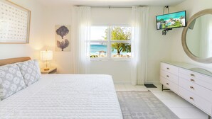 Een slaapkamer met een bed, nachtkastje, dressoir, televisie en een raam met uitzicht op het strand.