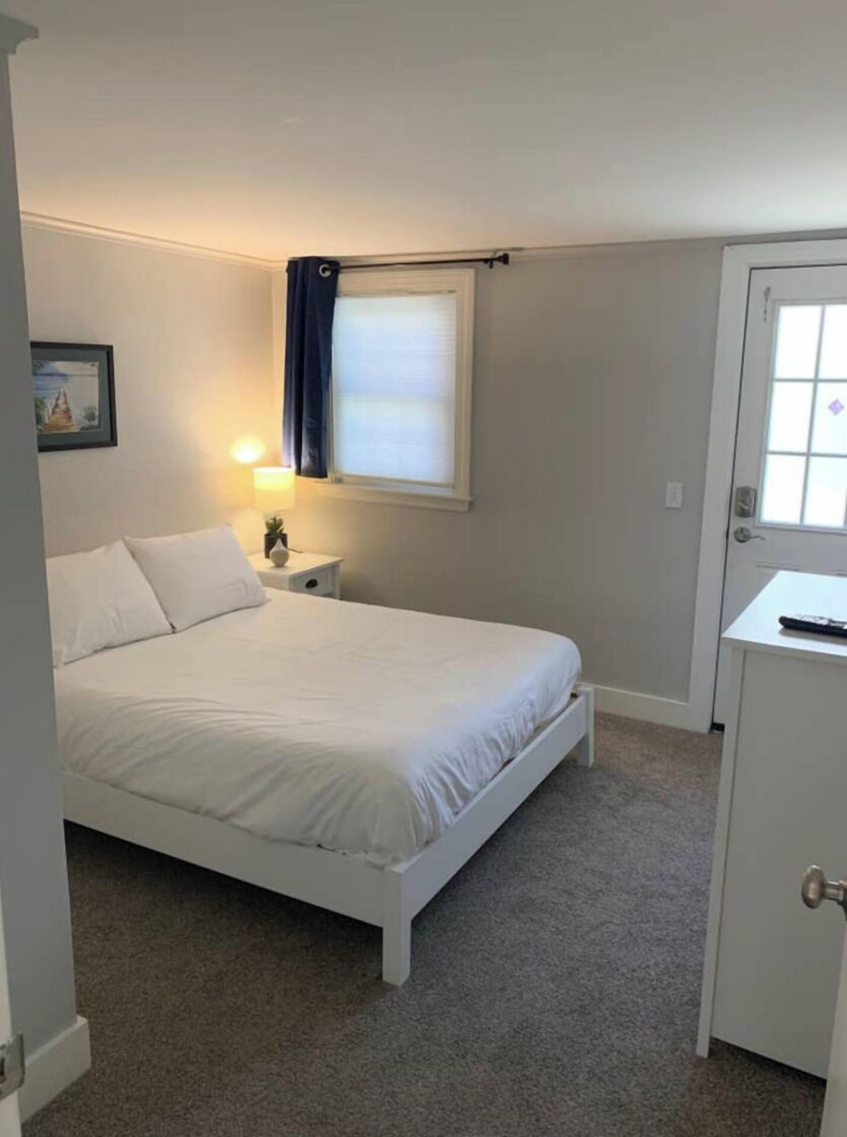 Een slaapkamer met een enkel bed, een nachtkastje, een lamp, een raam met gordijnen, een deur en een klein kastje.