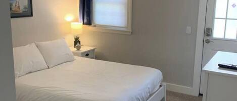 Een slaapkamer met een enkel bed, een nachtkastje, een lamp, een raam met gordijnen, een deur en een klein kastje.