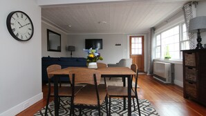 A dining area with a wooden table and chairs, a patterned rug, a blue sofa, a wall clock, and a television in the background. - 9 Lake Street  (Wolfeboro)
