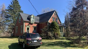 A two-story house with a red exterior and green trim, a car parked in the driveway, and a large tree in front.