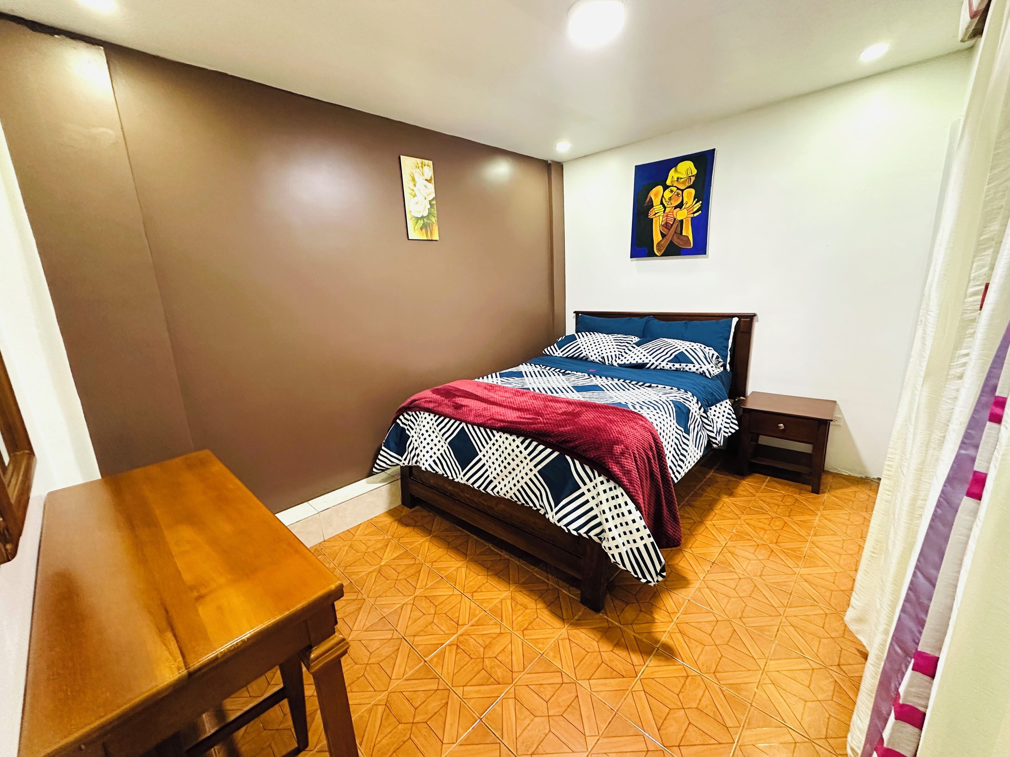 A bedroom with a bed, a wooden nightstand, a small table, and a wall with two paintings.