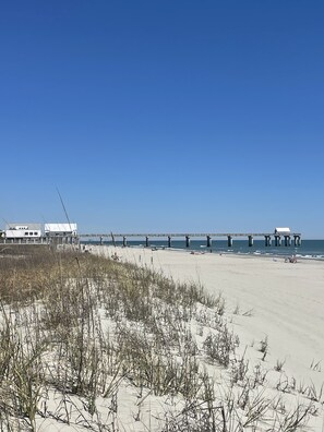 Een strand met een pier die in de oceaan uitloopt, zandduinen en gebouwen op de achtergrond.