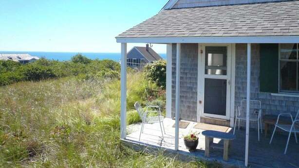 Una casa frente al mar con un porche cubierto, una puerta, una mesa pequeña y sillas.