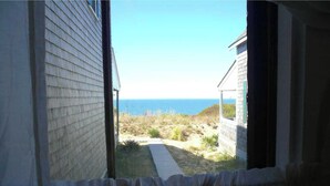 Vista desde una ventana sobre un sendero arenoso que lleva a una playa, con un edificio a la izquierda y una construcción a la derecha.
