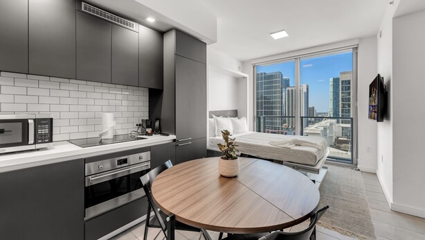 A modern kitchen with a dining area, a large window with a city view, and a bed with a cityscape view. - Stylish Studio + Den Across from Bayside Miami! (Miami)