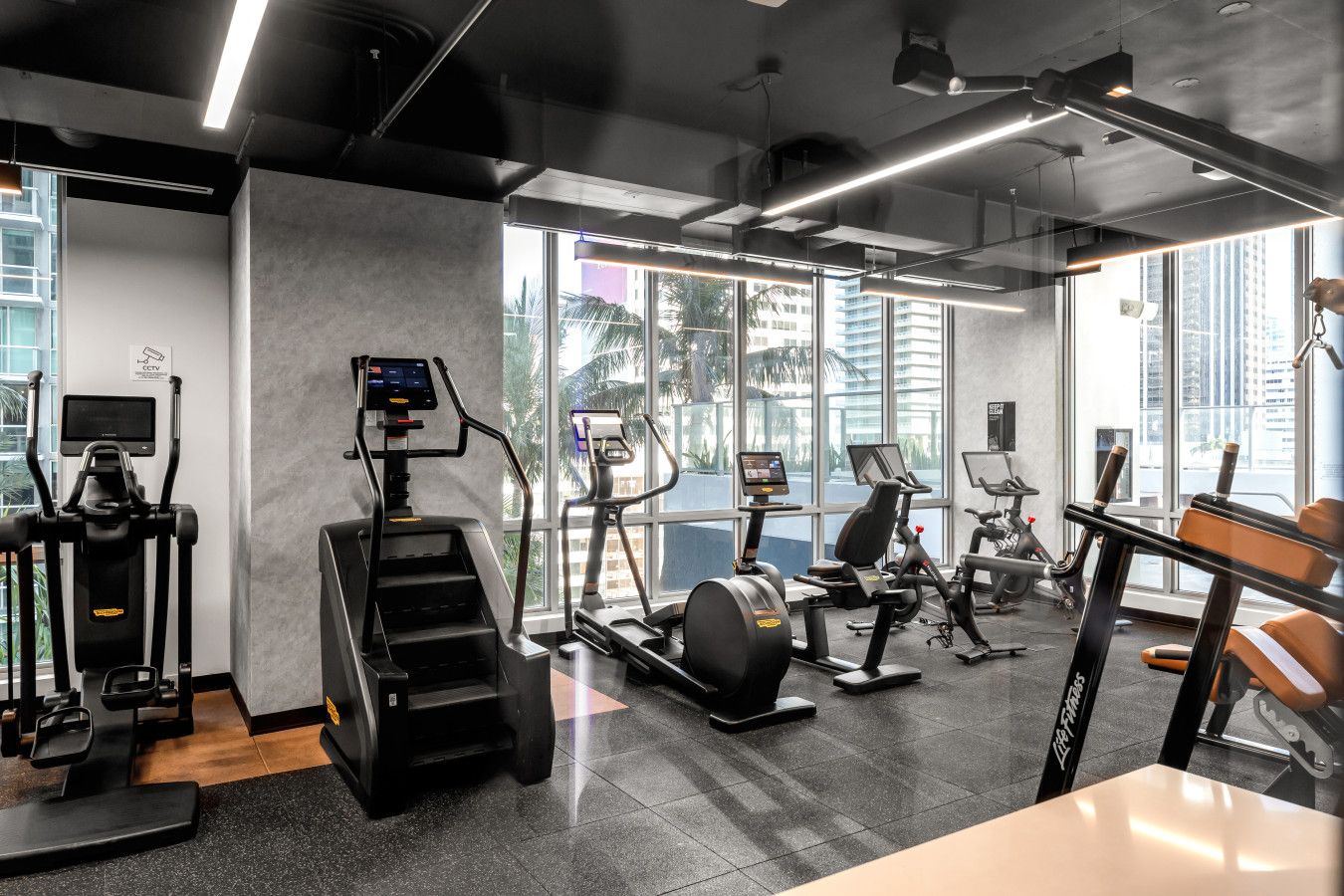 A modern gym with various cardio machines, a digital display, and large windows offering a view of the city.