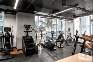 A modern gym with various cardio machines, a digital display, and large windows offering a view of the city.