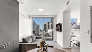 A modern apartment with a dining area, a grey sofa, and a view of the cityscape through large windows. - Stylish Studio + Den Across from Bayside Miami! (Miami)