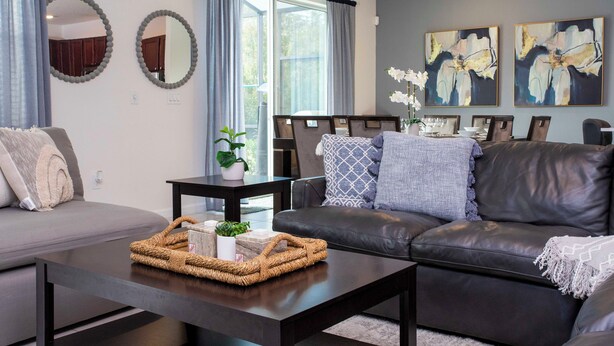 A modern living room with a grey sofa, a dark leather couch, a coffee table with a woven basket, and two abstract paintings on the wall. - Amazing 8BR Family Villa at Veranda Palms ● Private Pool/BBQ/More! (Kissimmee)
