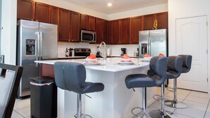 A modern kitchen with stainless steel appliances, wooden cabinets, and a central island with bar stools. - Amazing 8BR Family Villa at Veranda Palms ● Private Pool/BBQ/More! (Kissimmee)