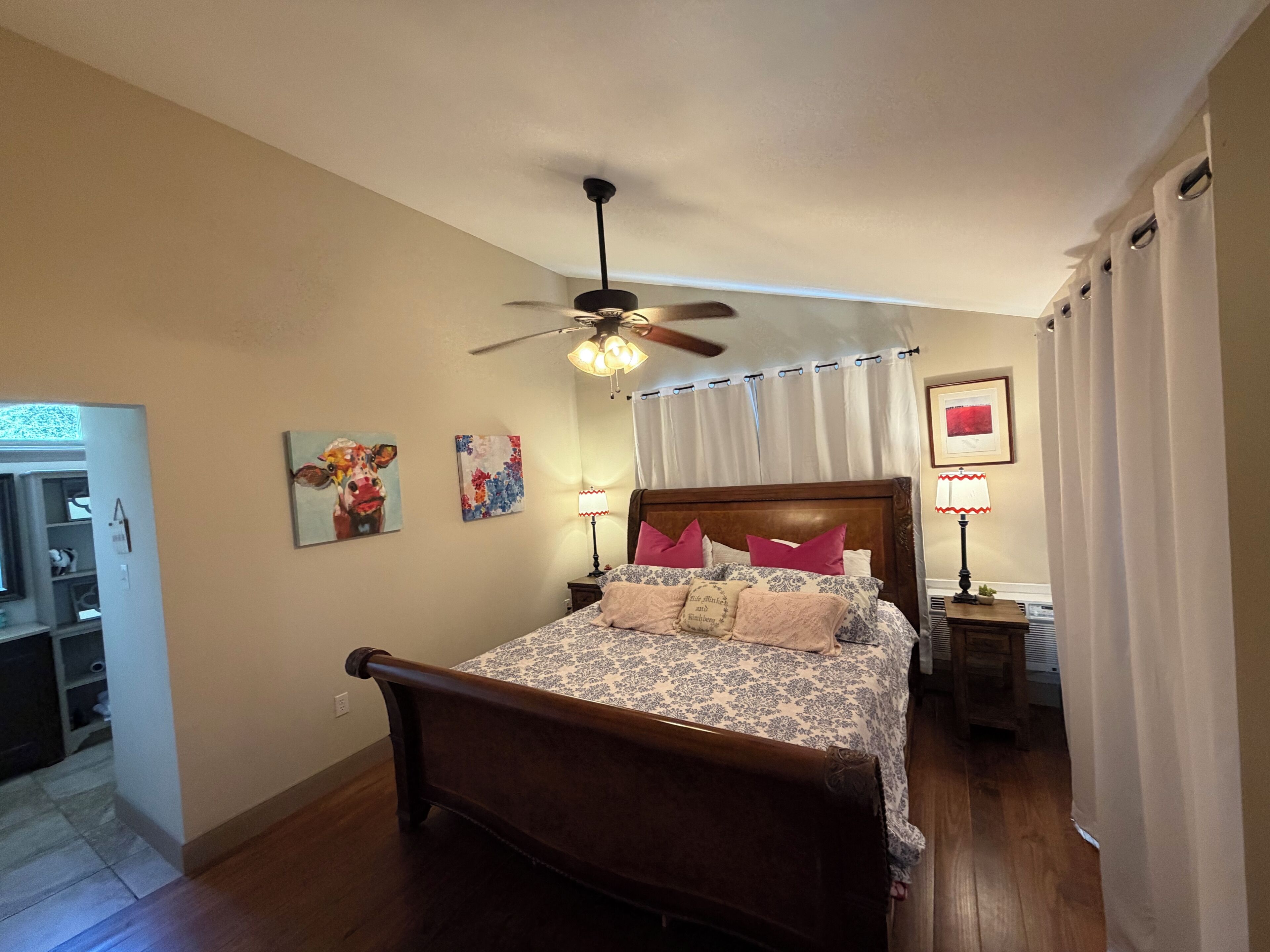 A bedroom with a wooden bed, a ceiling fan, a nightstand with a lamp, and a door leading to a bathroom.