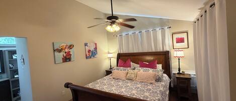 A bedroom with a wooden bed, a ceiling fan, a nightstand with a lamp, and a door leading to a bathroom.