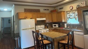 A kitchen with wooden cabinets, a refrigerator, stove, microwave, and a sink. There are two chairs at a table and a trash can. - 2-Br Country Cottage on rolling hills of Burton, 15 min from Round Top! (Burton)