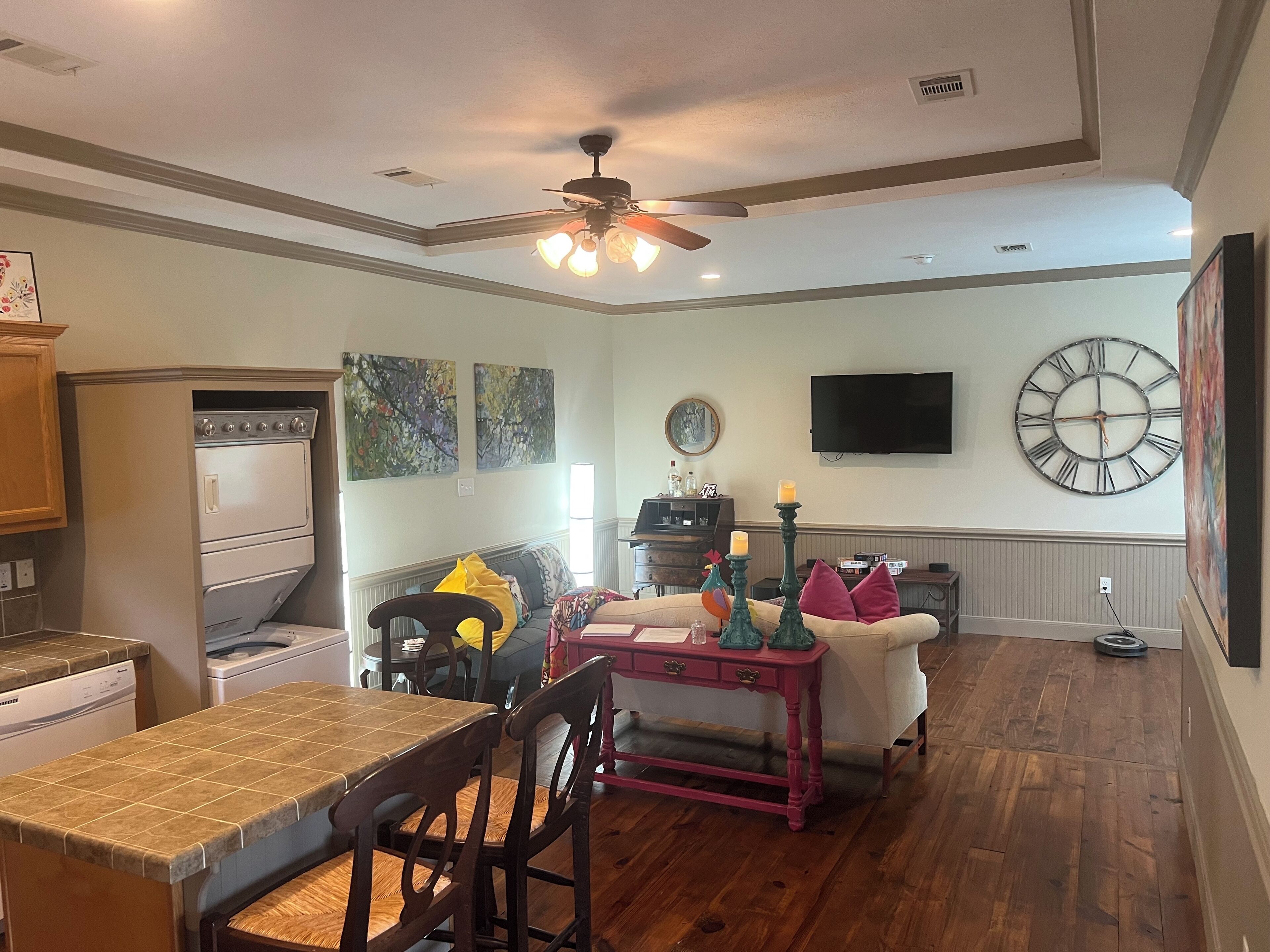A living room with a dining area, wooden flooring, a ceiling fan, a clock, and a TV on the wall.