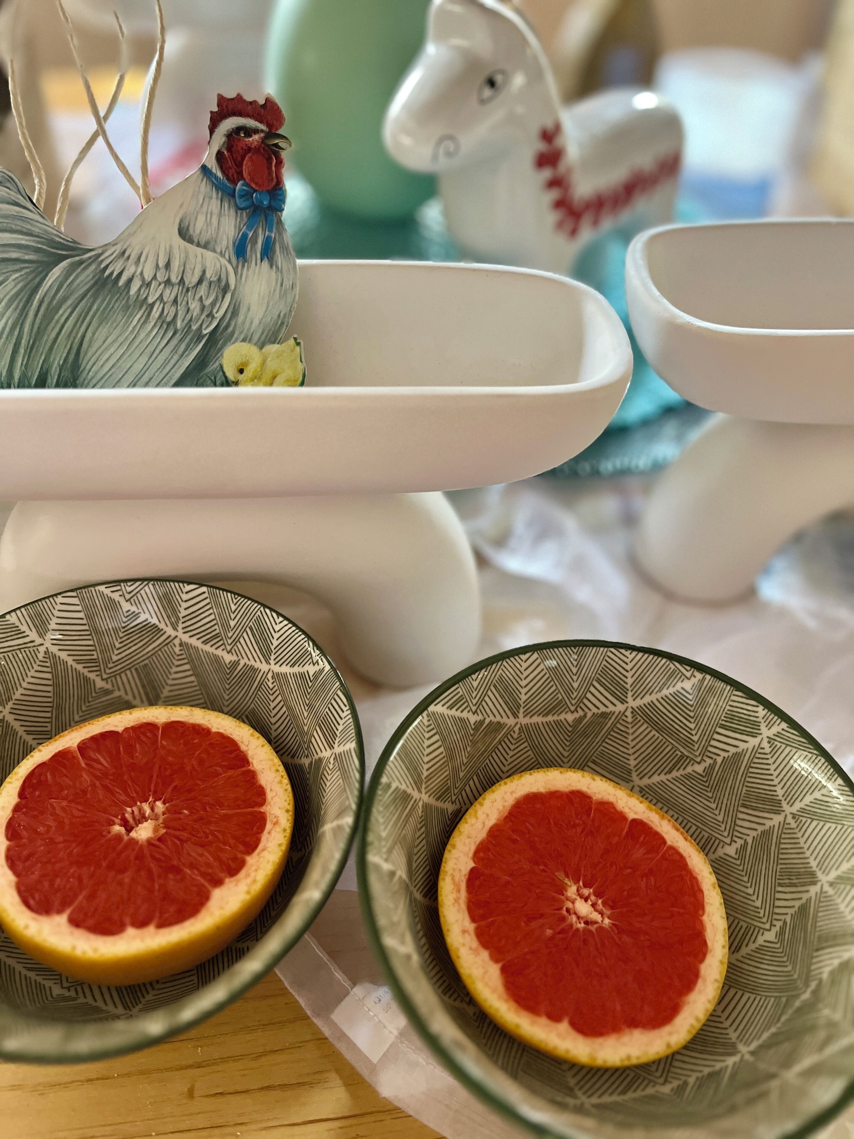 Une table sur laquelle reposent deux bols en verre décoré contenant des moitiés de pamplemousse, un coq en céramique et une figurine de cheval en céramique.
