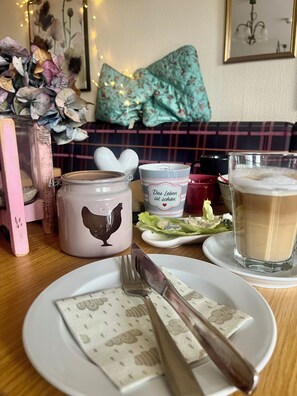 A dining table with a plate of food, a glass of beer, and a mug with a chicken design. - B&B Apart Hotel Goldener Hahn-Guesthouse (Baiersbronn)