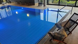 Una piscina interior moderna con baldosas azules cristalinas, una zona de terraza de madera con sillones reclinables y vistas a la vegetación a través de amplias ventanas.