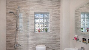 A bathroom with a glass shower enclosure, a toilet, a sink, and a window with decorative items. - M City Apartments | Garden 2 | 100m from the beach (Chania)