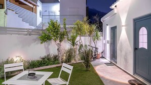 A modern outdoor patio with a table and chairs, surrounded by greenery and a white wall. - M City Apartments | Garden 2 | 100m from the beach (Chania)