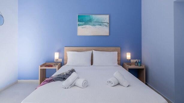 A hotel room with a large bed, two bedside tables, a painting of a beach scene, and a blue wall. - M City Apartments | Garden 2 | 100m from the beach (Chania)