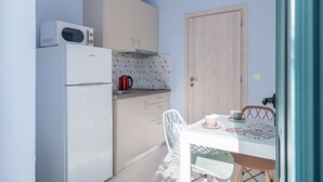 A compact kitchen with a refrigerator, microwave, and kettle, a wooden door, and a dining table with chairs. - M City Apartments | Garden 1 | 100m from the beach (Chania)