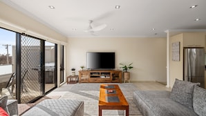 A living room with a flat-screen TV, wooden entertainment unit, sofa, and a sliding glass door leading to an outdoor area. - Brooklya by the Beach (Cowes)
