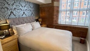 A bedroom with a large bed, a headboard with a patterned design, two bedside tables with lamps, and a window with a view of buildings. - Cocoa Suites-2 Bed-city Centre-pets-parking (York)