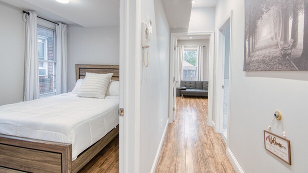 A bedroom with a wooden bed, a white wall, and a hallway leading to another room.
